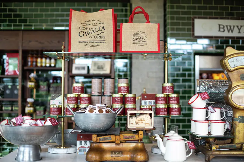 A shop display featuring branded hessian bags, tins of goods, enamel mugs, and a vintage brass scale against green tiles.