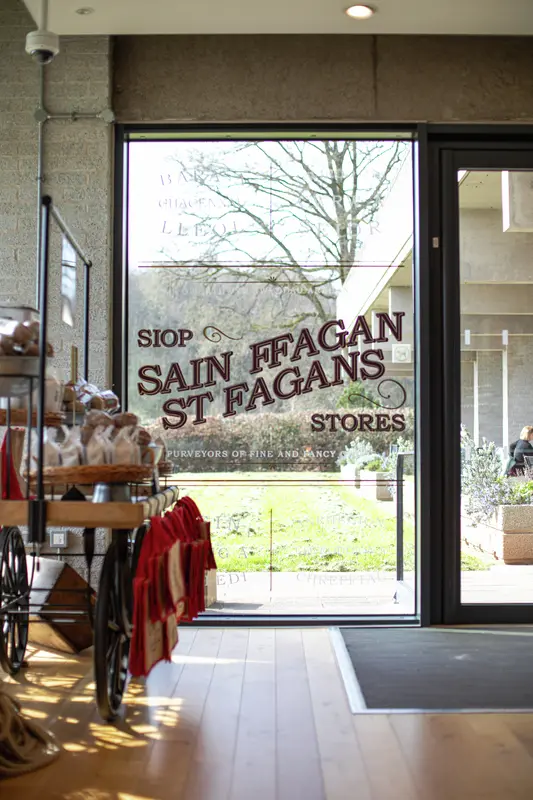 A shop window with 'St Fagans Stores' lettering, looking out onto a sunny garden with a wooden cart in the foreground.