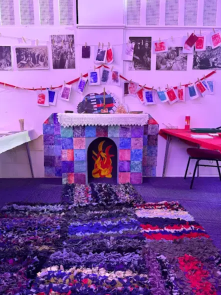 Colourful fireplace created with cardboard, rag mat on the floor and hanging prints and family portraits on wall with a few artefacts on mantle piece creating a living room experience.