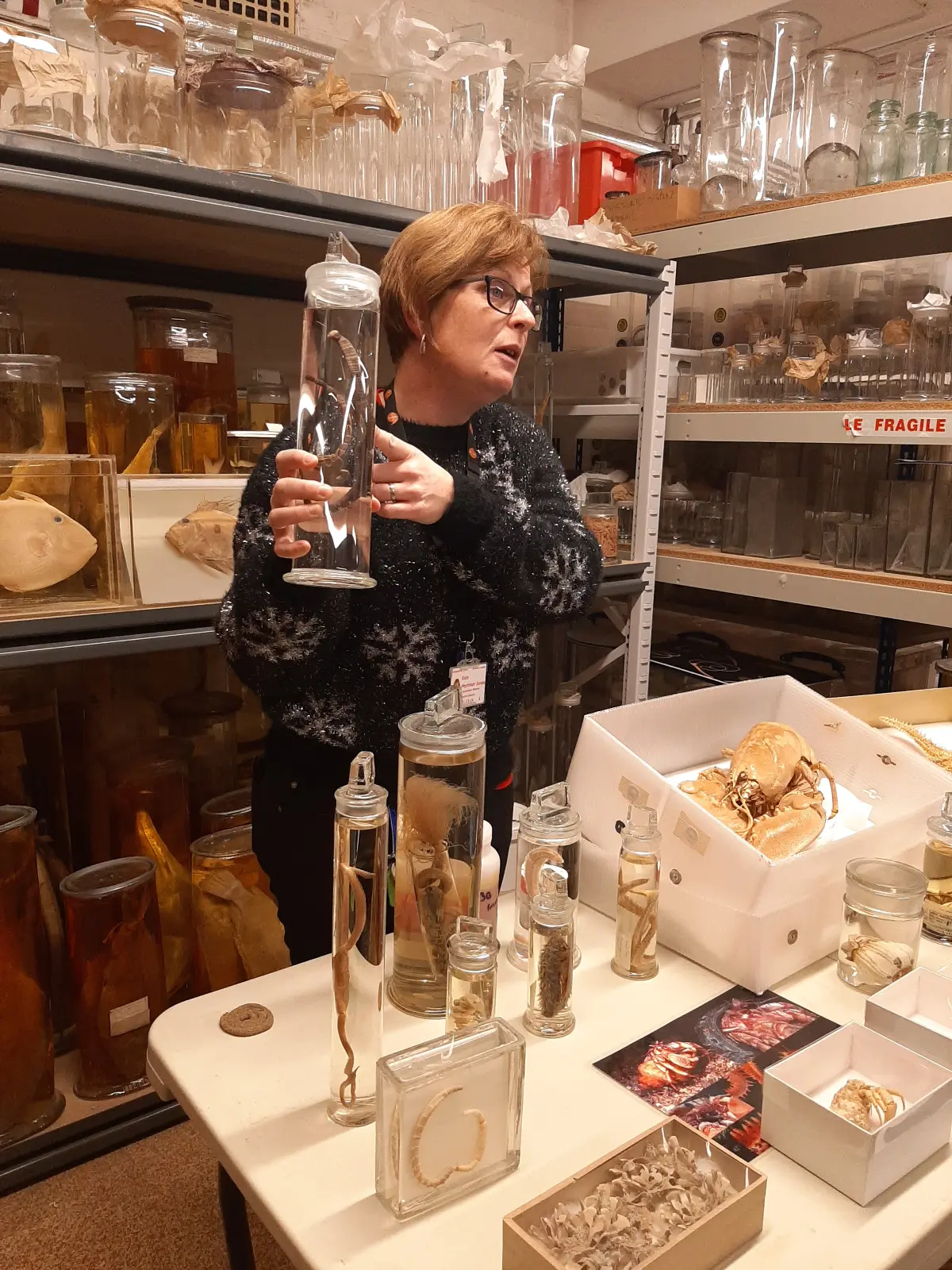 A woman is wearing a black jumper with white snowflakes over it, she is surrounded by speciments and flasks while holding a jar herself